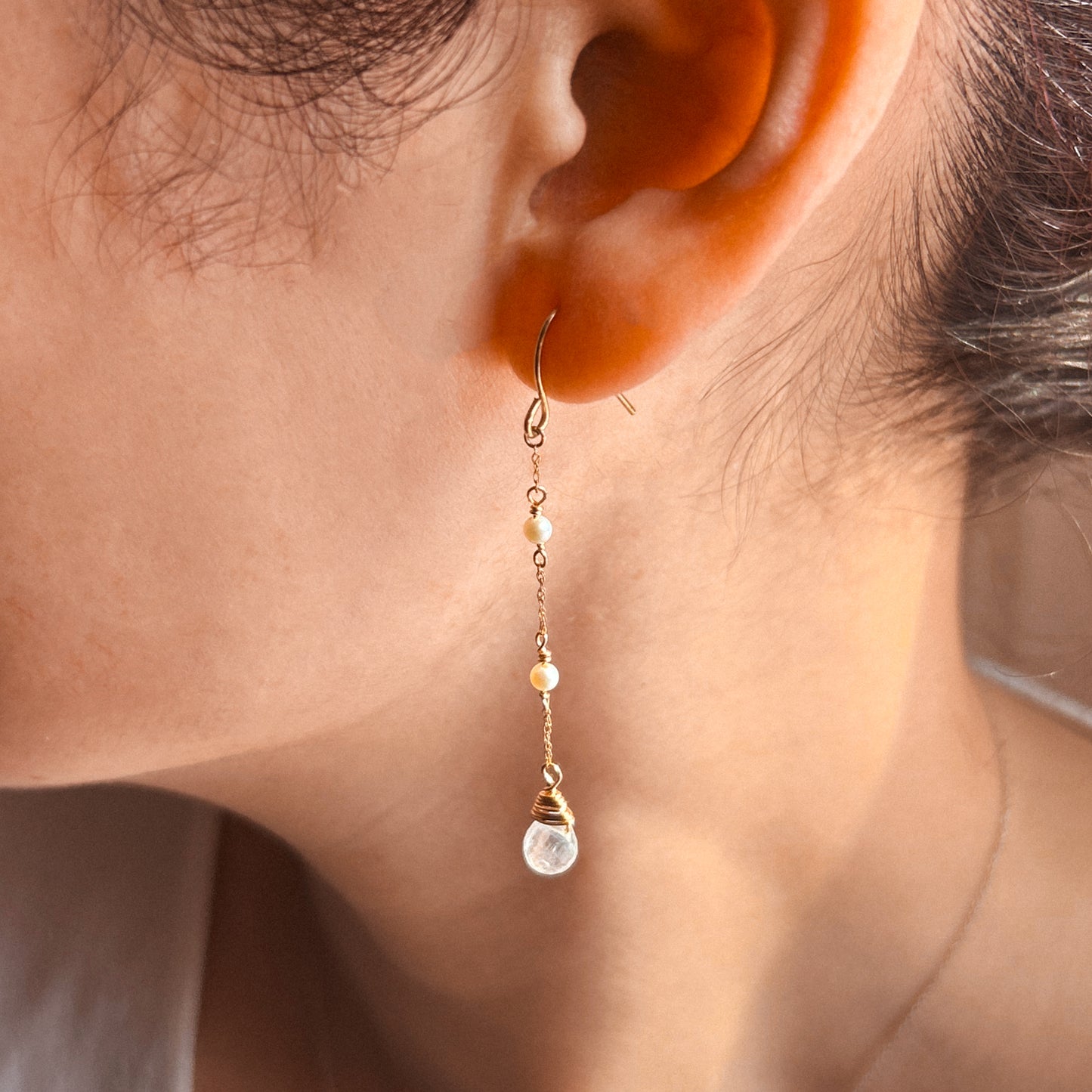 A pair of gold drop earrings featuring Akoya pearls and moonstone, with the earrings suspended against a white background.