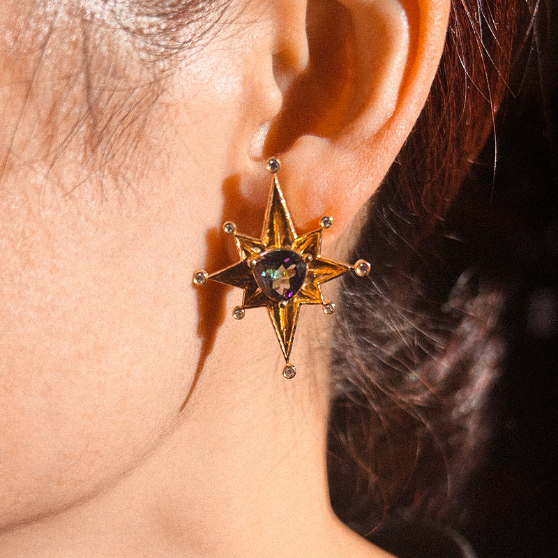 A pair of gold-colored earrings with star-shaped designs and purple Mystic Tourmaline at the center.