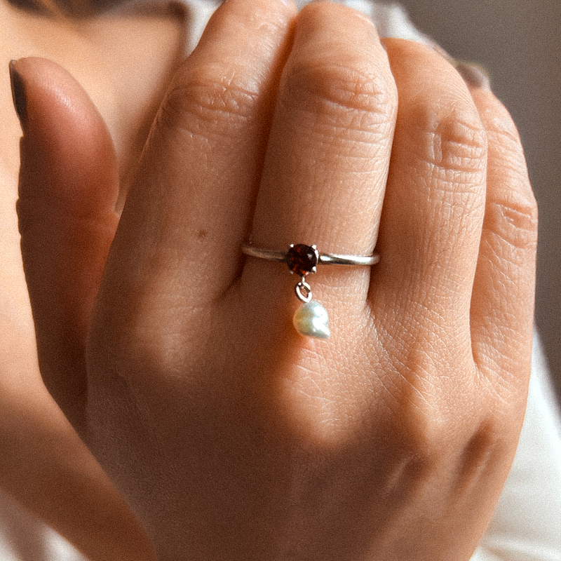 Close-up of a hand wearing a silver ring with a red gemstone and pearl charm.