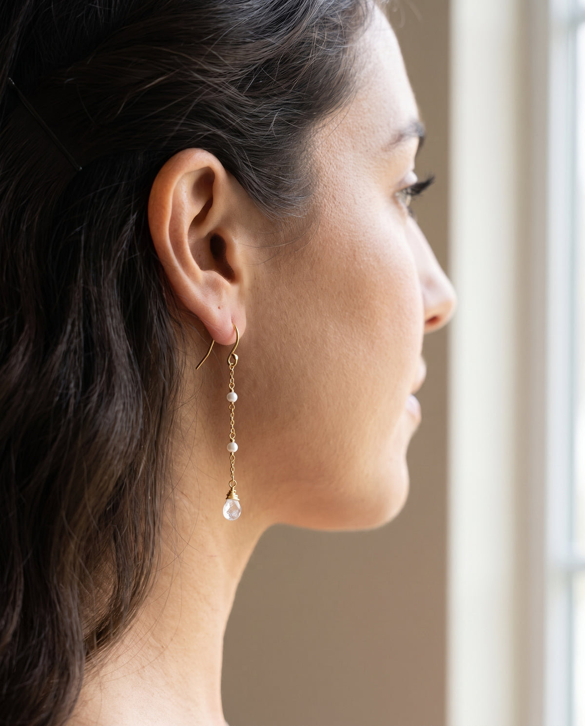 A close-up side profile of a woman wearing a delicate gold dangle with moonstone earring with small white pearl-like beads and a teardrop moonstone drop, set against a soft beige background near a window.