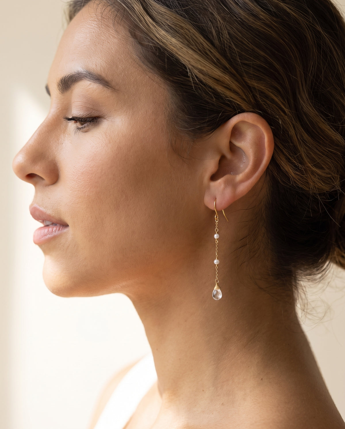 A close-up side profile of a woman wearing a delicate gold dangle with moonstone earring with small clear stones and a teardrop-shaped clear moonstone drop, set against a soft, warm neutral background.