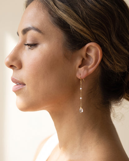 A close-up side profile of a woman wearing a delicate gold dangle with moonstone earring with small clear stones and a teardrop-shaped clear moonstone drop, set against a soft, warm neutral background.